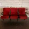 Three red upholstered theater seats with black metal arms sit in a row against a worn white brick wall.