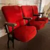 Row of three red upholstered theater seats with metal armrests against a rough concrete wall in an industrial space.