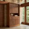 Wooden cabinet with vertical ribbed doors set against a stone wall and large windows showing greenery outside