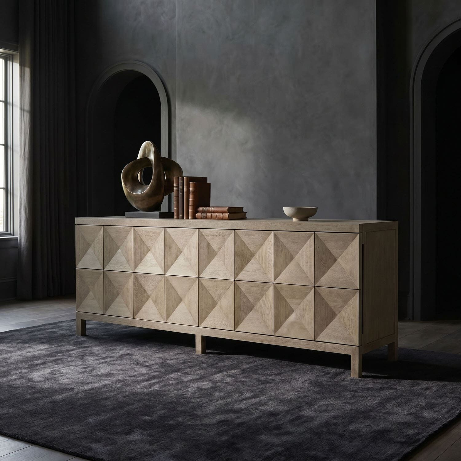 Beige wooden sideboard with a geometric diamond pattern, topped by a bronze abstract sculpture, a row of books, and a small bowl in a dim, modern living room.