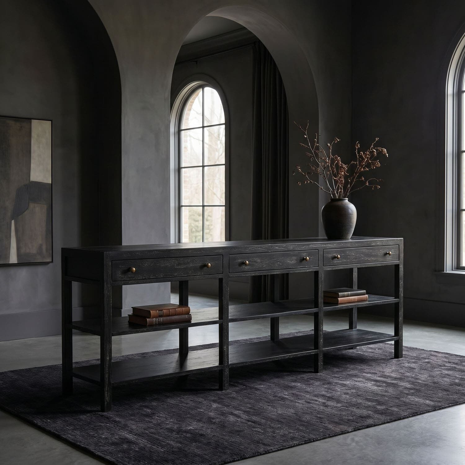 Moody, minimalist room with a black wooden console table, a dark vase with bare branches, and arched windows in the background on a dark rug.