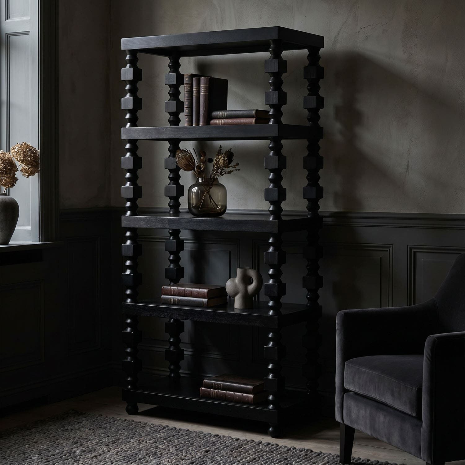 Dark, sculpted wooden bookshelf with stacked spindled columns holding books and a glass vase of dried flowers in a moody, dimly lit room.